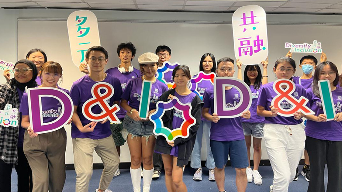 A group of D&I ambassadors stands together on a stage, smiling and holding props at the D&I Ambassador Programme Opening Ceremony.