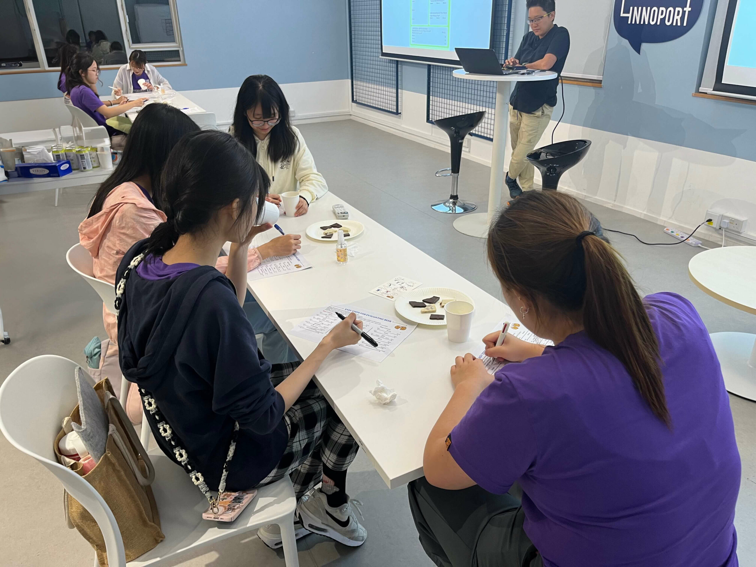 Students seated at tables in a classroom, writing, while a teacher presents at the front during the ‘Beer, Chocolate & Inclusion’ workshop.