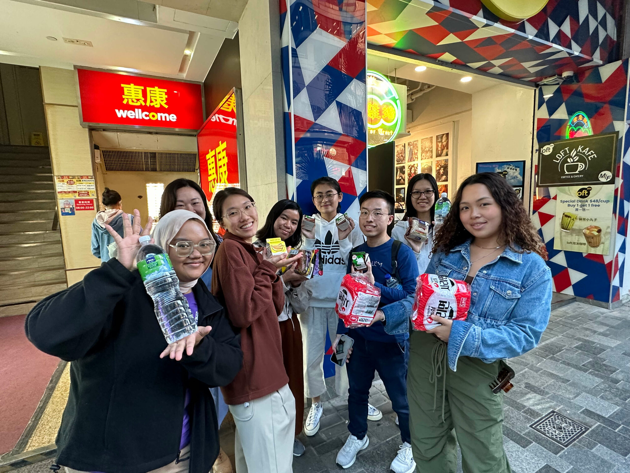 The ambassadors standing outside a Wellcome store holding various items like snacks and drinks.