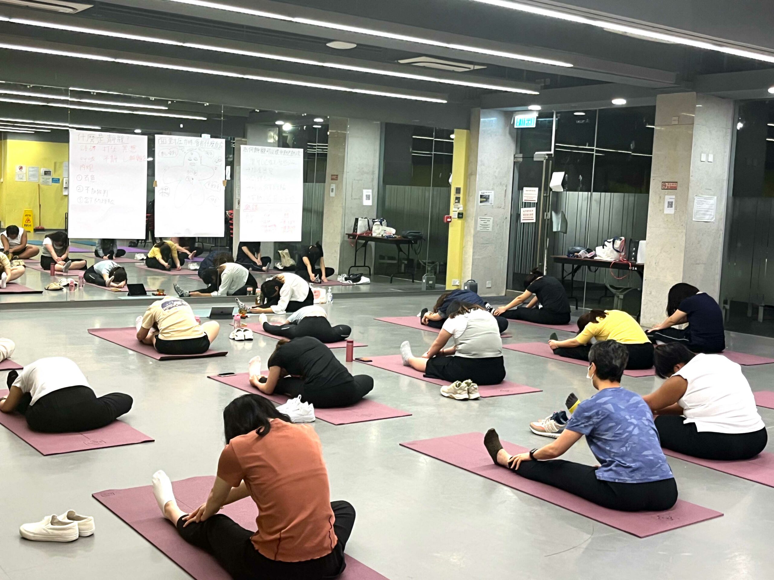 A group of people practicing yoga in a mirrored studio, seated on pink mats, doing forward bends during a class.