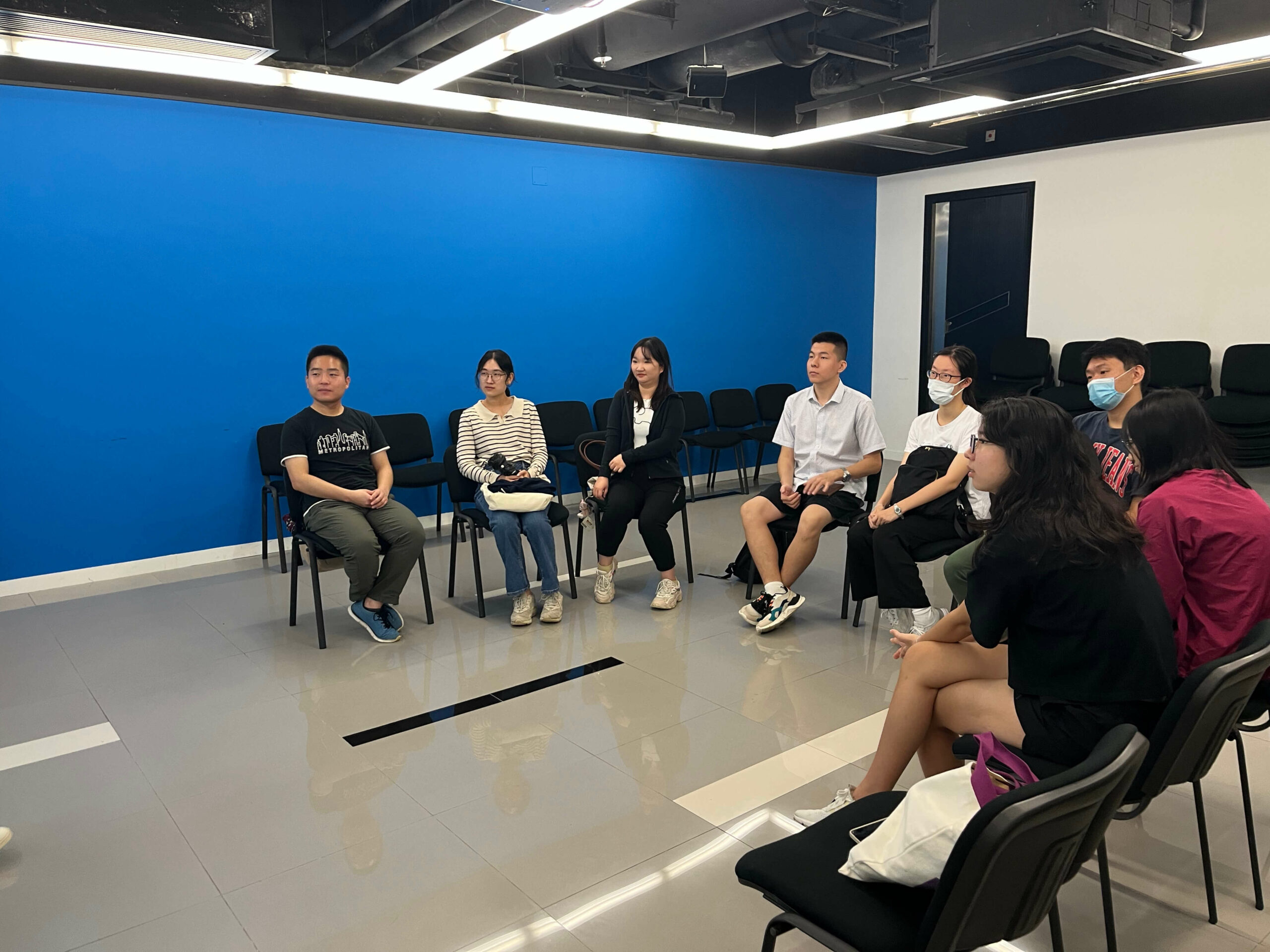 A group of people sitting in a circle of chairs in a room with a blue wall and a gray floor, participating in a discussion before the city hunt event.