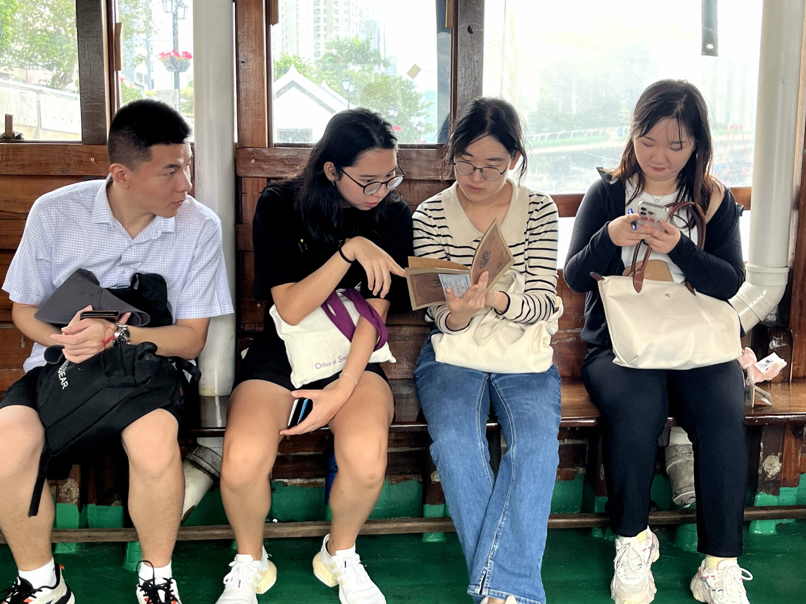 this Four people sit on a wooden bench outdoors or in a semi-outdoor setting with windows showing greenery and buildings. One holds a black bag, one holds a phone, one reads a book, and the last looks at a phone.