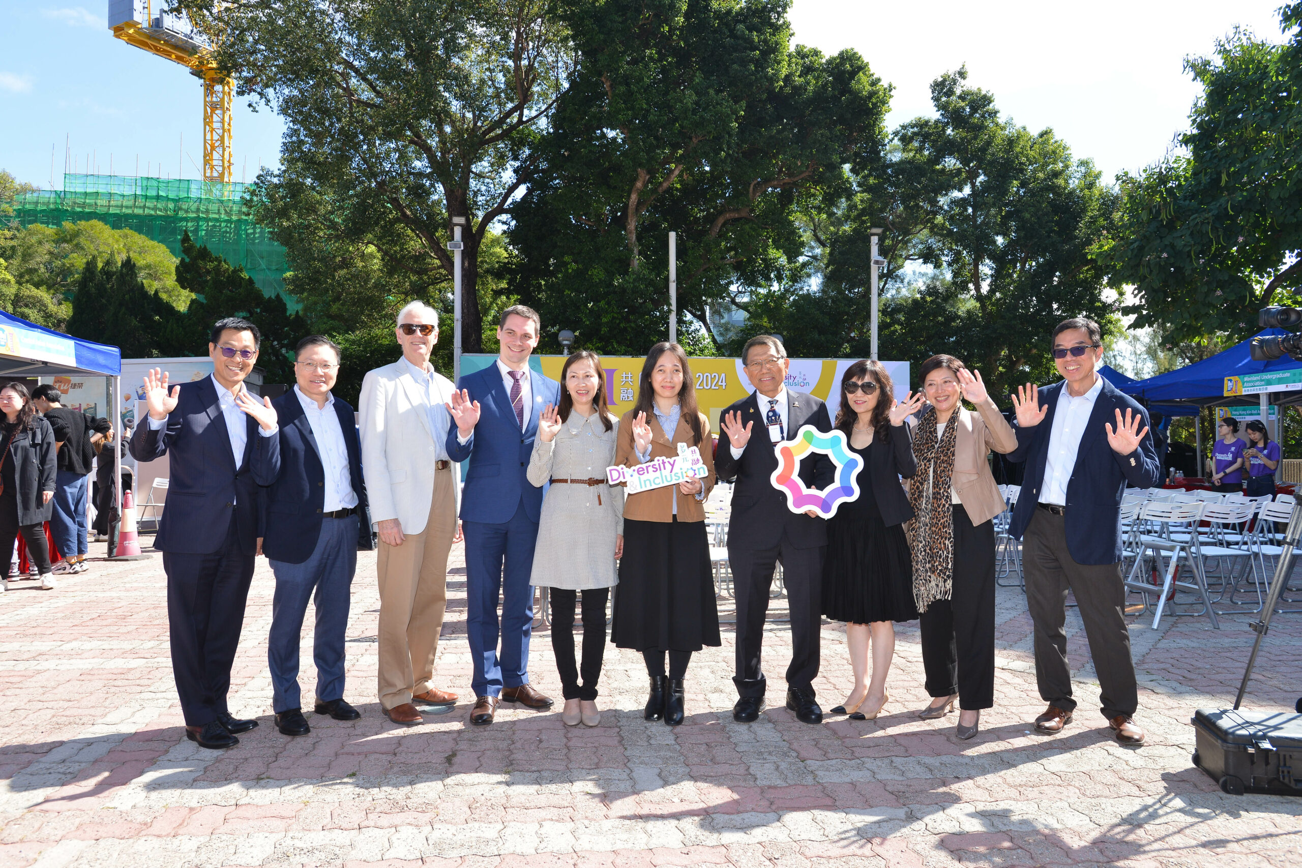 Nine guests, inlcuding the Vice-Chancellor of CUHK, Rocky Tuan, stand outdoors in a line, posing for a photo. They hold "Diversity & Inclusion" signs, with trees, a crane, and tents in the background.