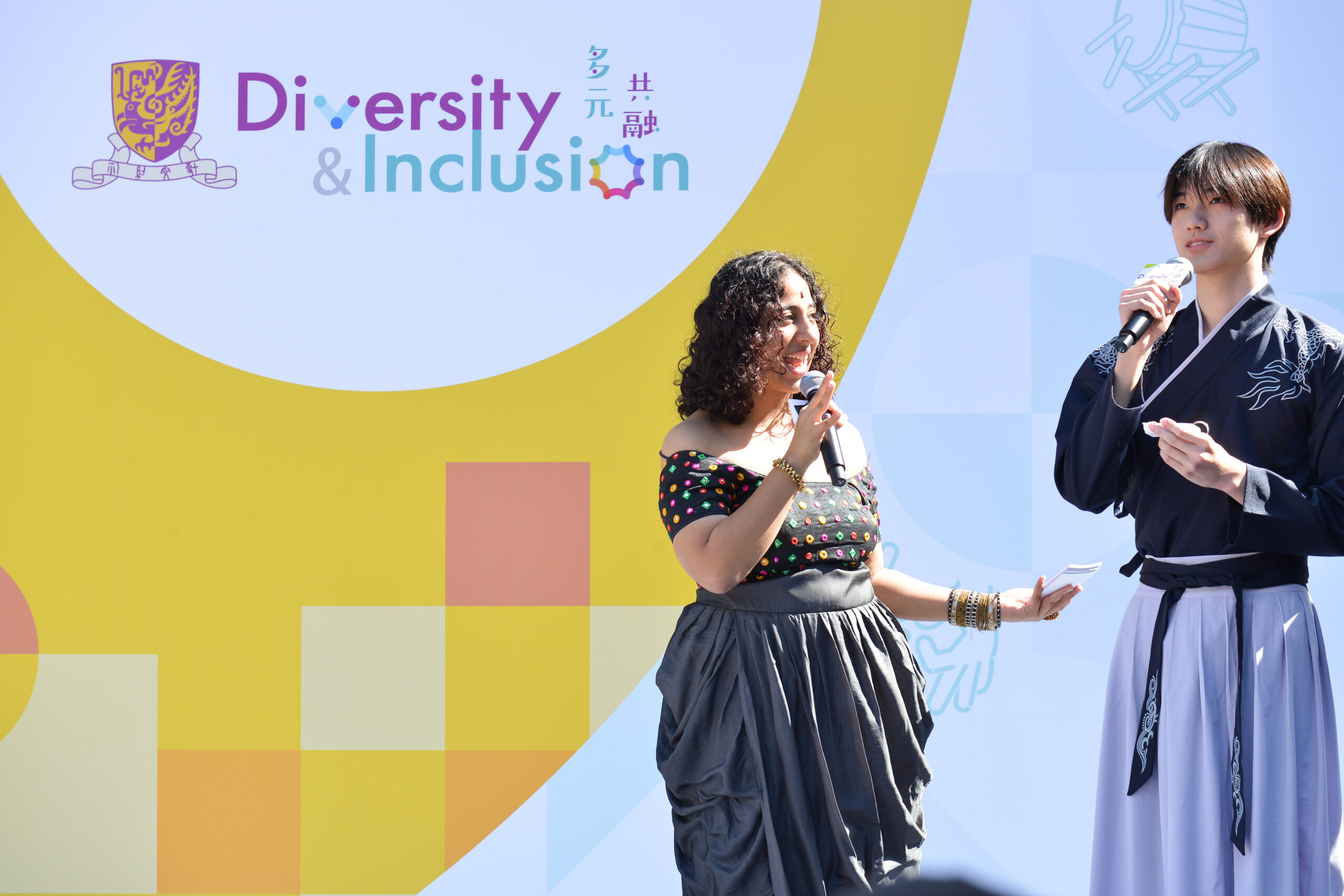 Two MCs on stage holding microphones, with a "Diversity & Inclusion" backdrop in English and Chinese behind them. One wears a colorful top, the other traditional attire.