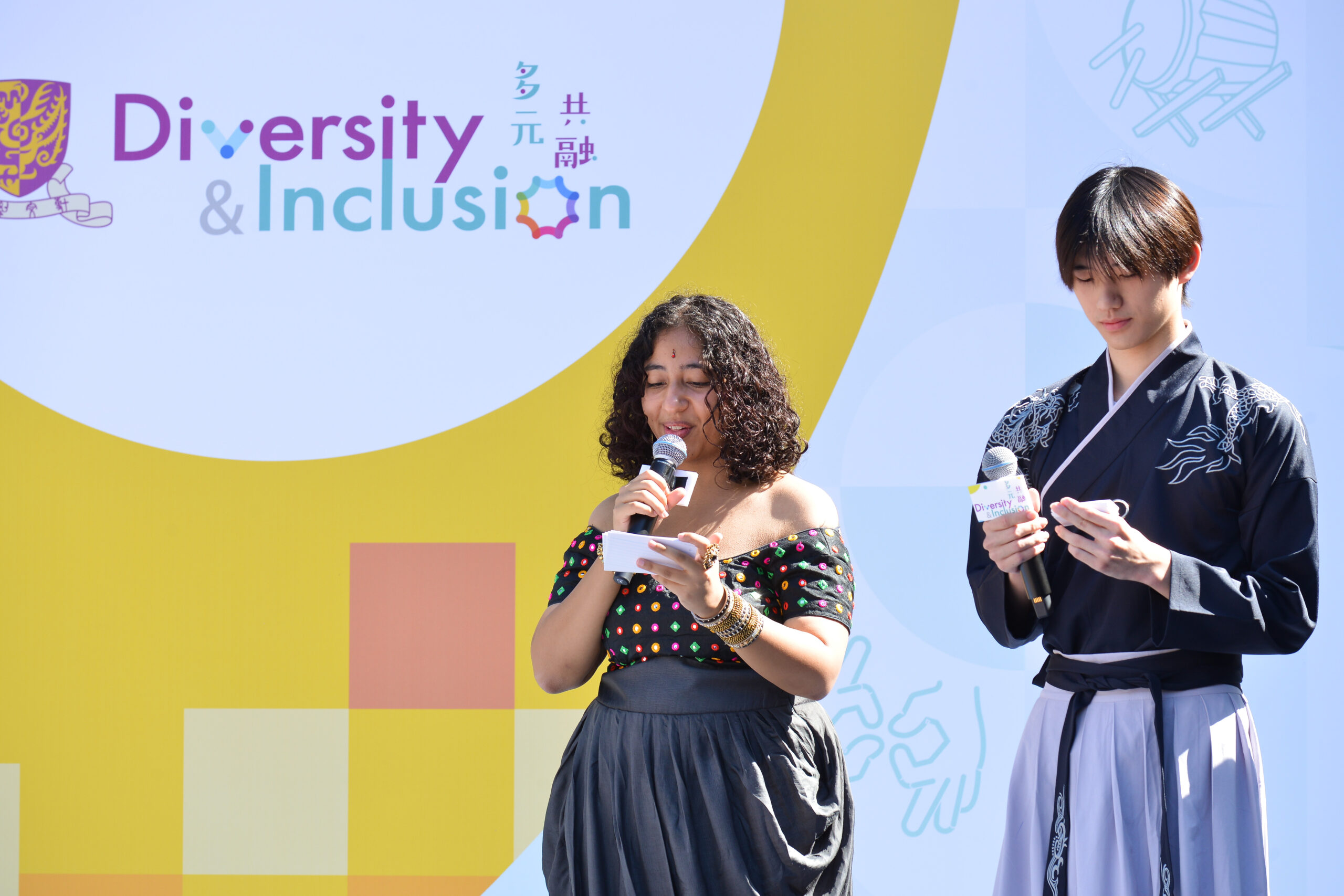 Two MCs on stage holding microphones, with a "Diversity & Inclusion" backdrop in English and Chinese behind them. One wears a colorful top, the other traditional attire.