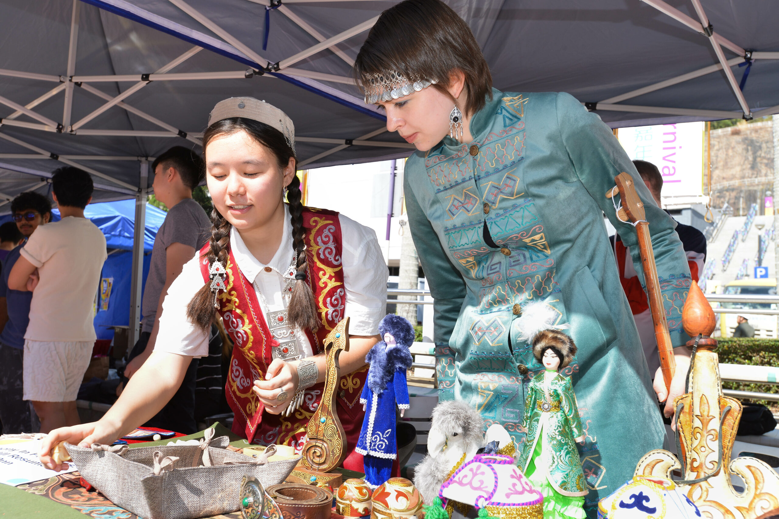 Two students from Central Asian Student Association in traditional clothing displaying handcrafted items, including dolls, carvings, and decorative objects.