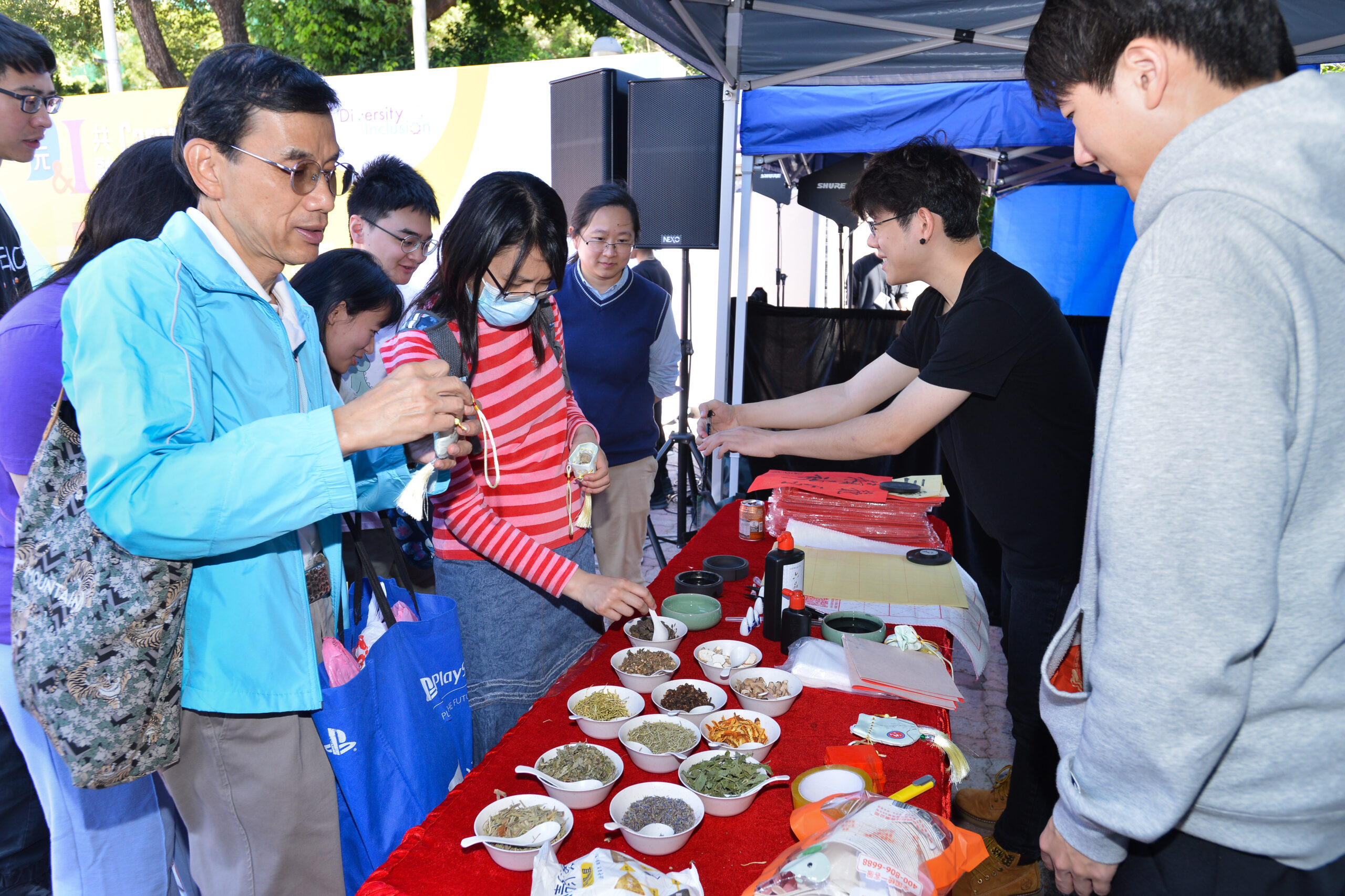 Participants at D&I Carnival 2024 interacting at an outdoor booth with a variety of herbs, spices, and other items on a red-covered table