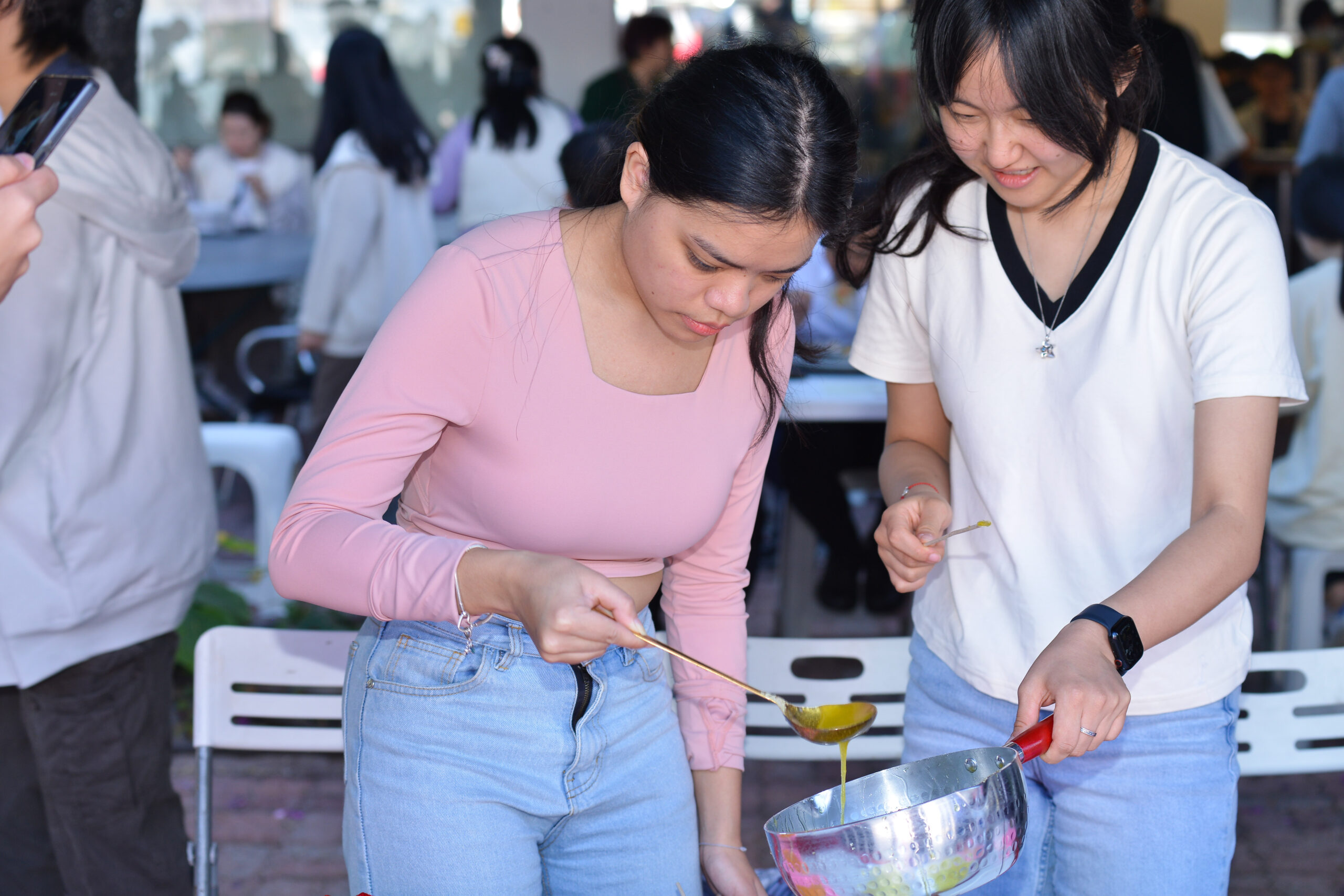 Two students at D&I Carnival 2024, collaborating to pour the liquid sugar from a metal bowl in the workshop.