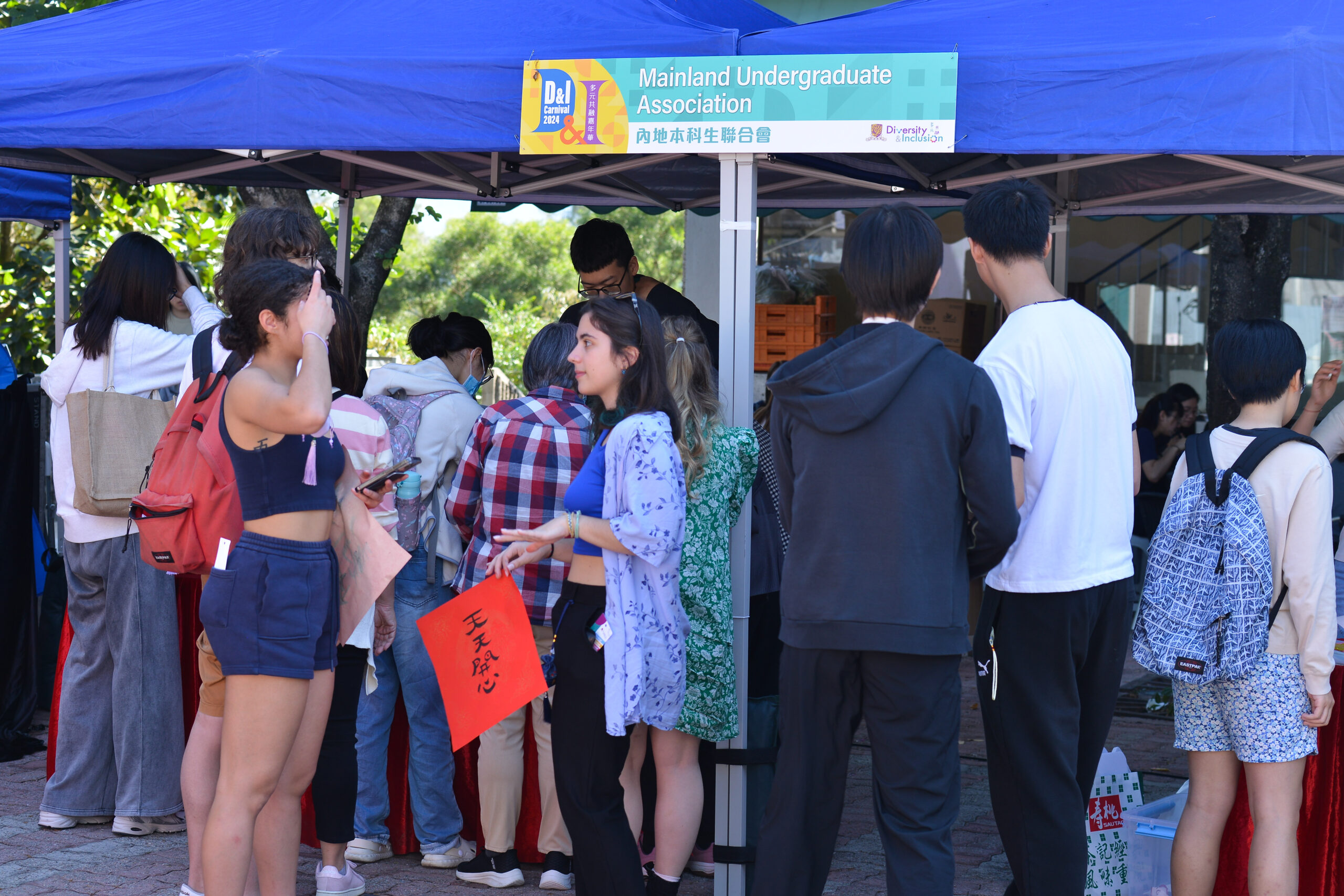 A group of students under a blue tent, engaging in conversation at D&I Carnival 2024, with one person holding fai chun.