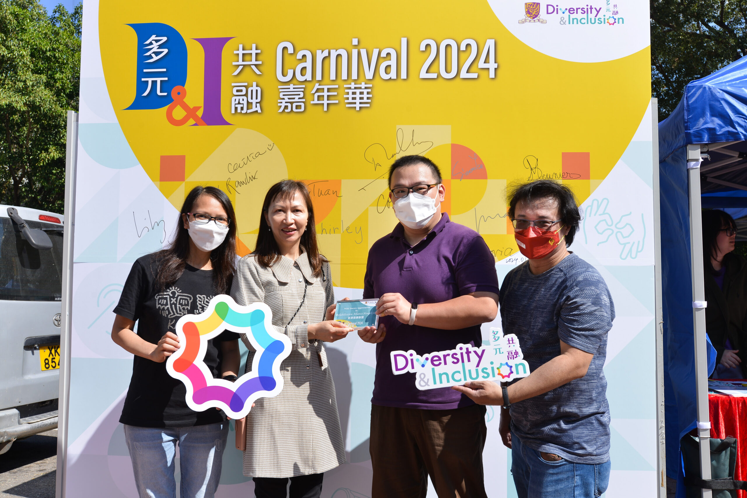 Four participants standing in front of a colorful backdrop at D&I Carnival 2024, holding signs and a plaque promoting diversity and inclusion.