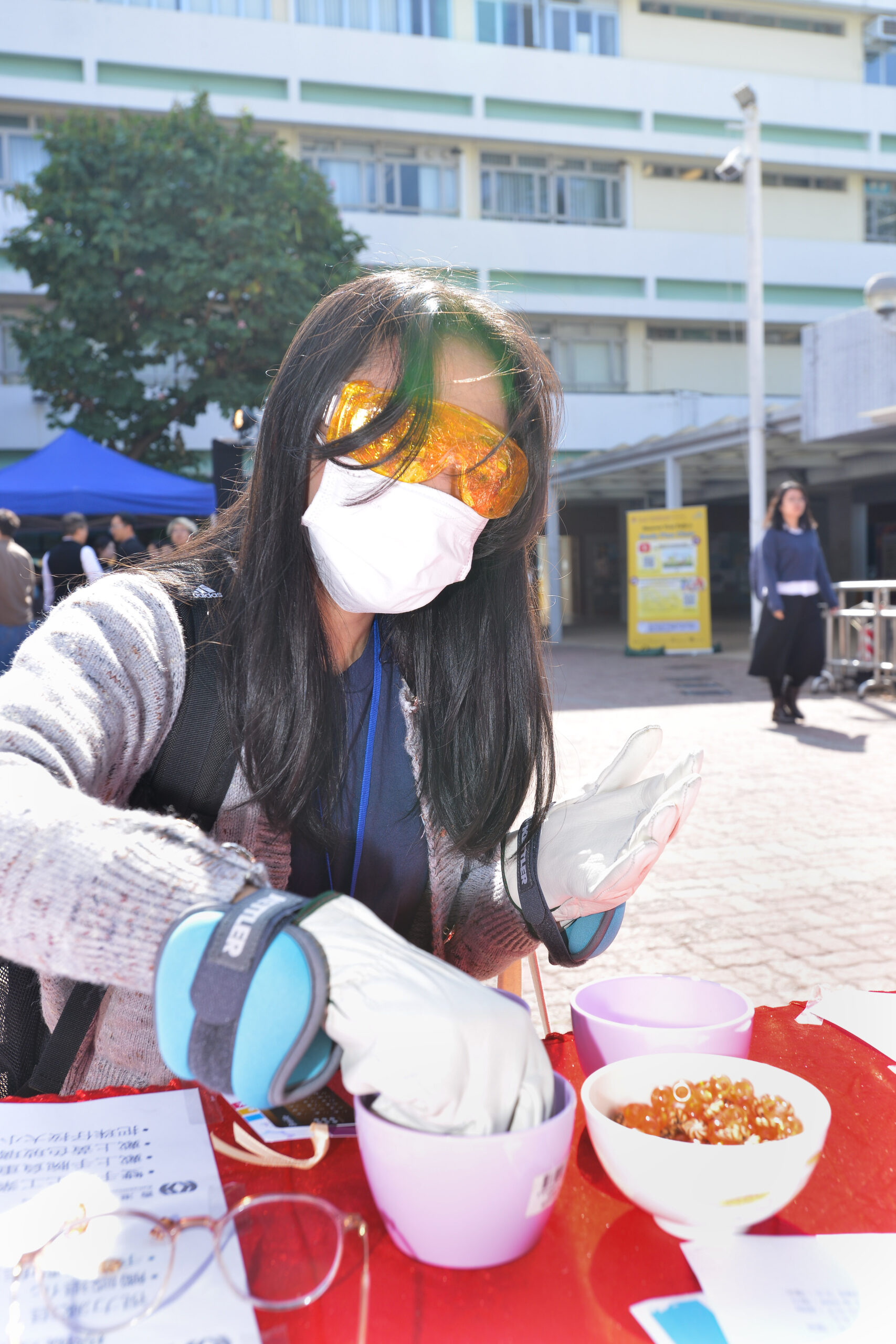 Person participating in an outdoor activity at D&I Carnival 2024, seated at a red table with bowls of beads and papers.