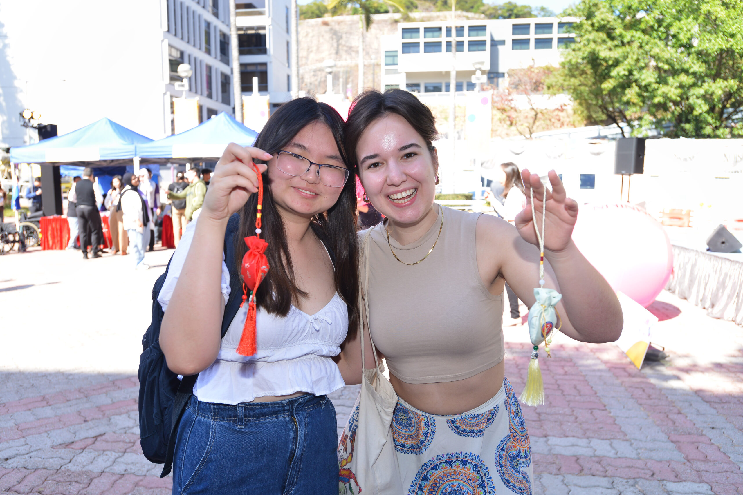 Two participants at D&I Carnival 2024 holding decorative pouches with tassels, standing outdoors at the event.