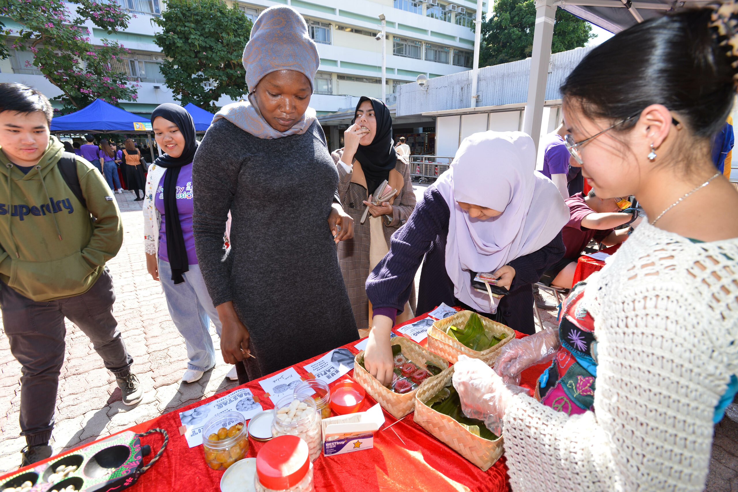 Group at a table covered with a red cloth at the carnival, displaying and engaging with various items like baskets with leaves, jars, and containers