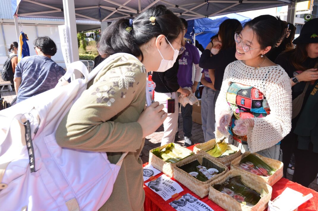 "Participants at D&I Carnival 2024 gathered around a food stall displaying traditional items like 'LUPIS GULUNG' and 'KLEPON'."