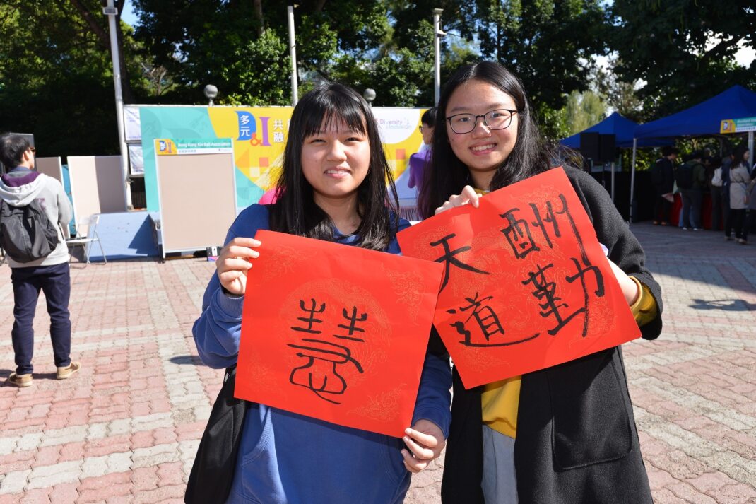 Two students holding the fai chun they wrote at D&I Carnival 2024, with booths and other participants in the background outdoors.