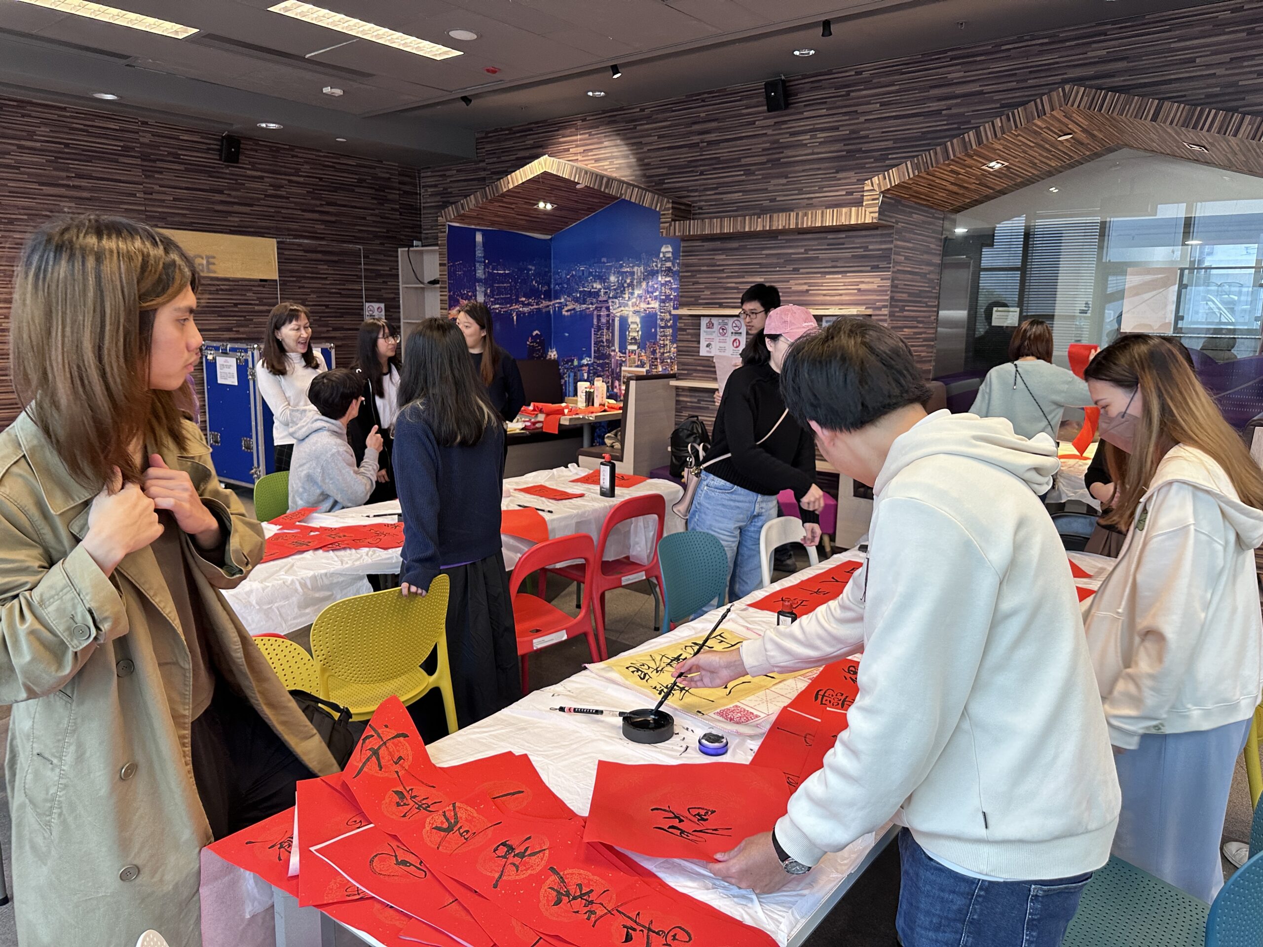 A person practices Chinese calligraphy at a Fai Chun workshop, writing characters on red paper with a brush.