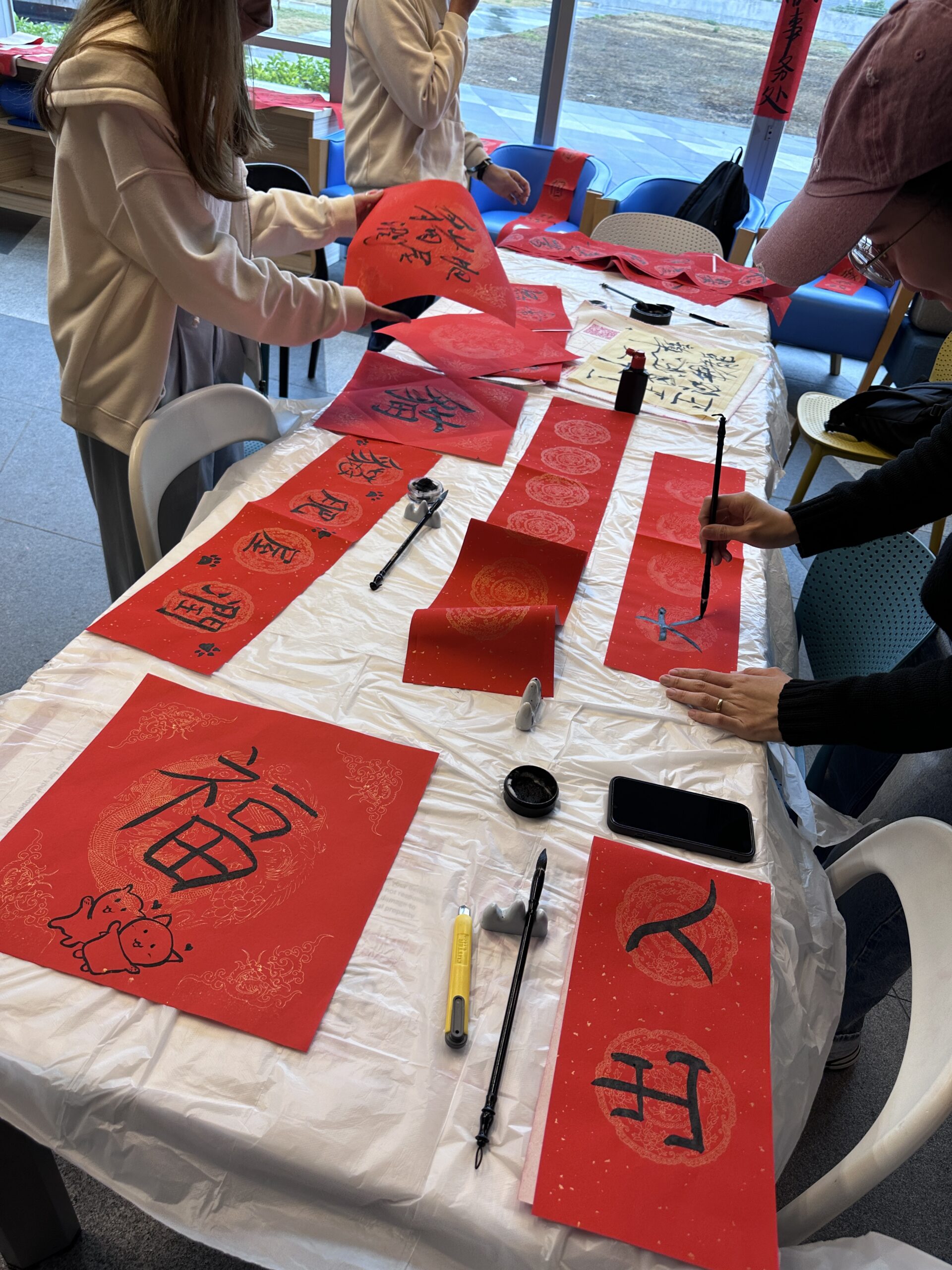 Five people sit at a table during a Fai Chun workshop. They are engaged in writing Chinese characters on red paper, using brushes and ink