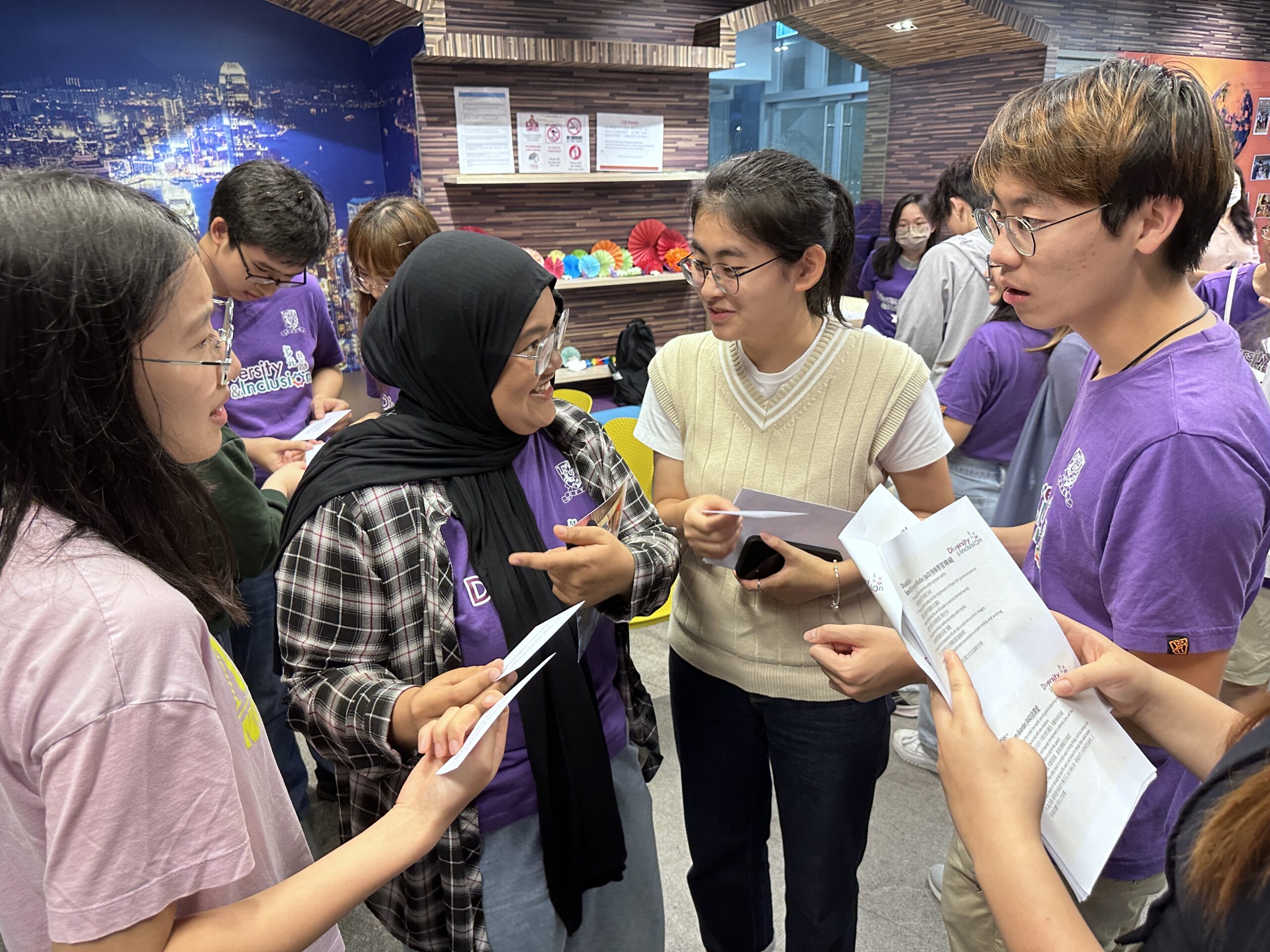 A group of people wearing purple "Diversity & Inclusion" shirts engage in happy conversation at the D&I Ambassador Programme Closing Ceremony, holding papers.