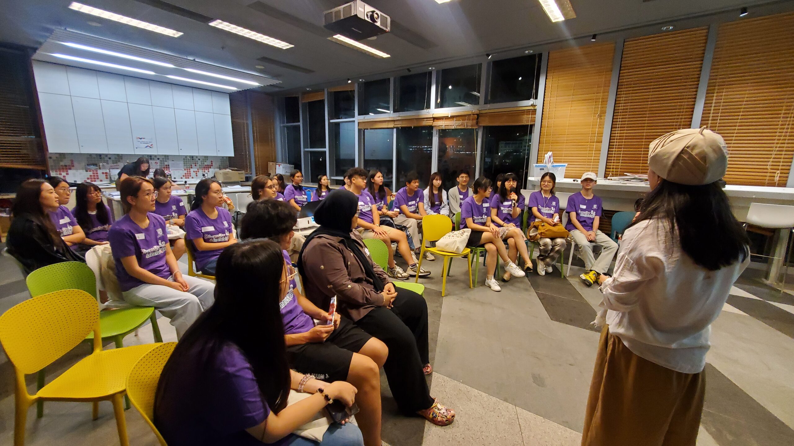 D&I ambassadors are seated in a semi-circle in a room with large windows and blinds. Most wear purple shirts, and a speker stands in front, presenting to them.