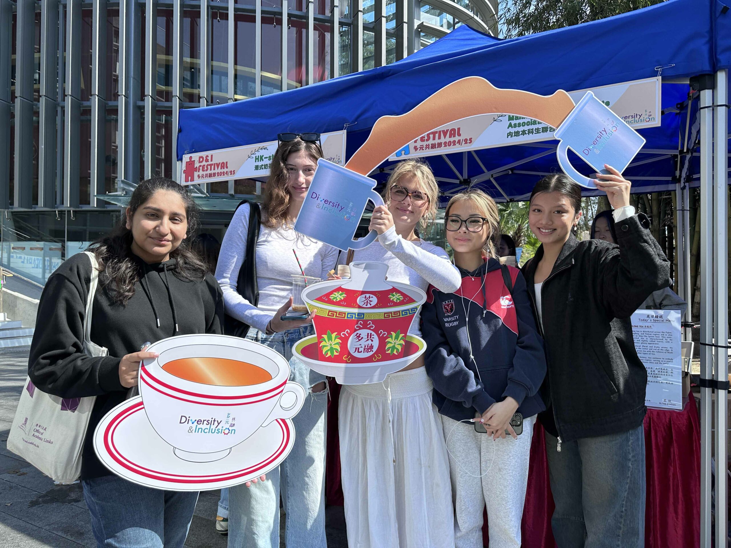 Five students are taking a photo with tea-like props and smiling in front of the event booth.