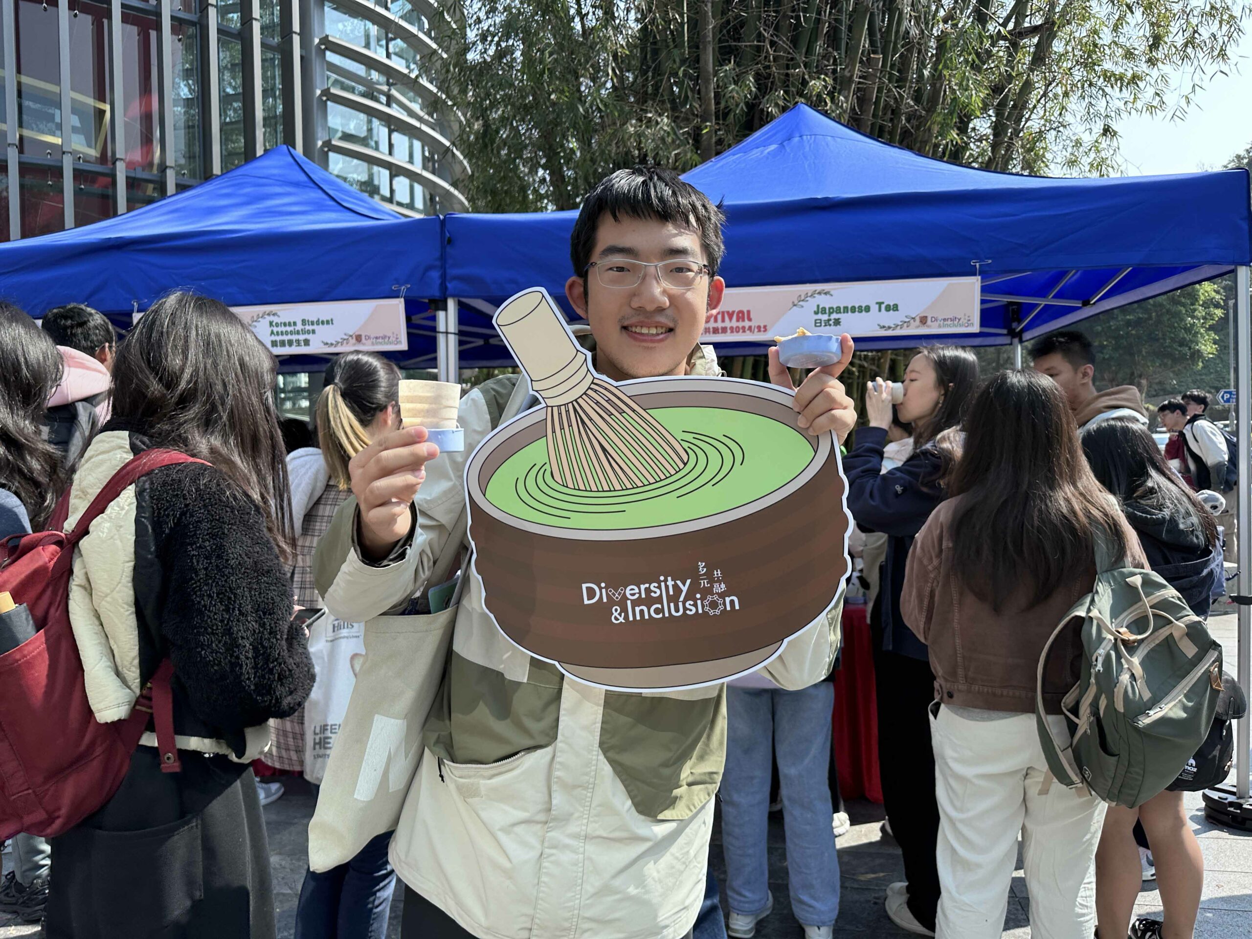 A student is taking a photo with a “matcha” prop and a blue foldable cup in front of the event booth.