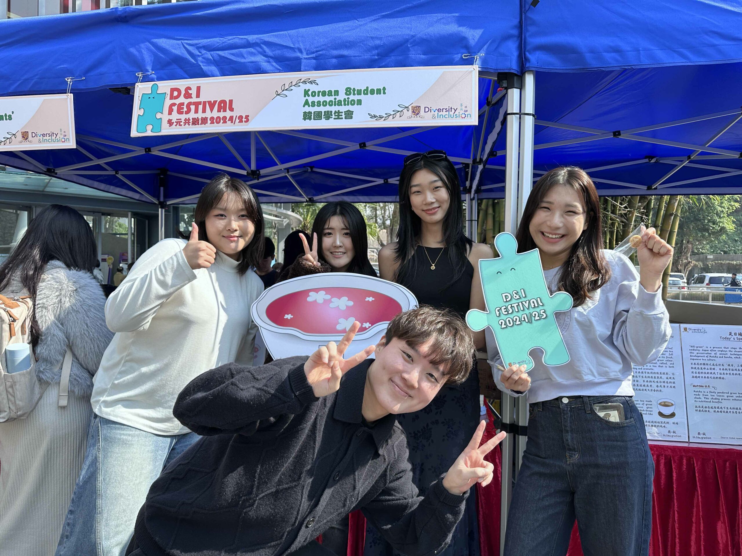 Five students from the Korean Student Association are smiling and taking photos with “omija tea” props under a blue tent with a "D&I Festival" banner.