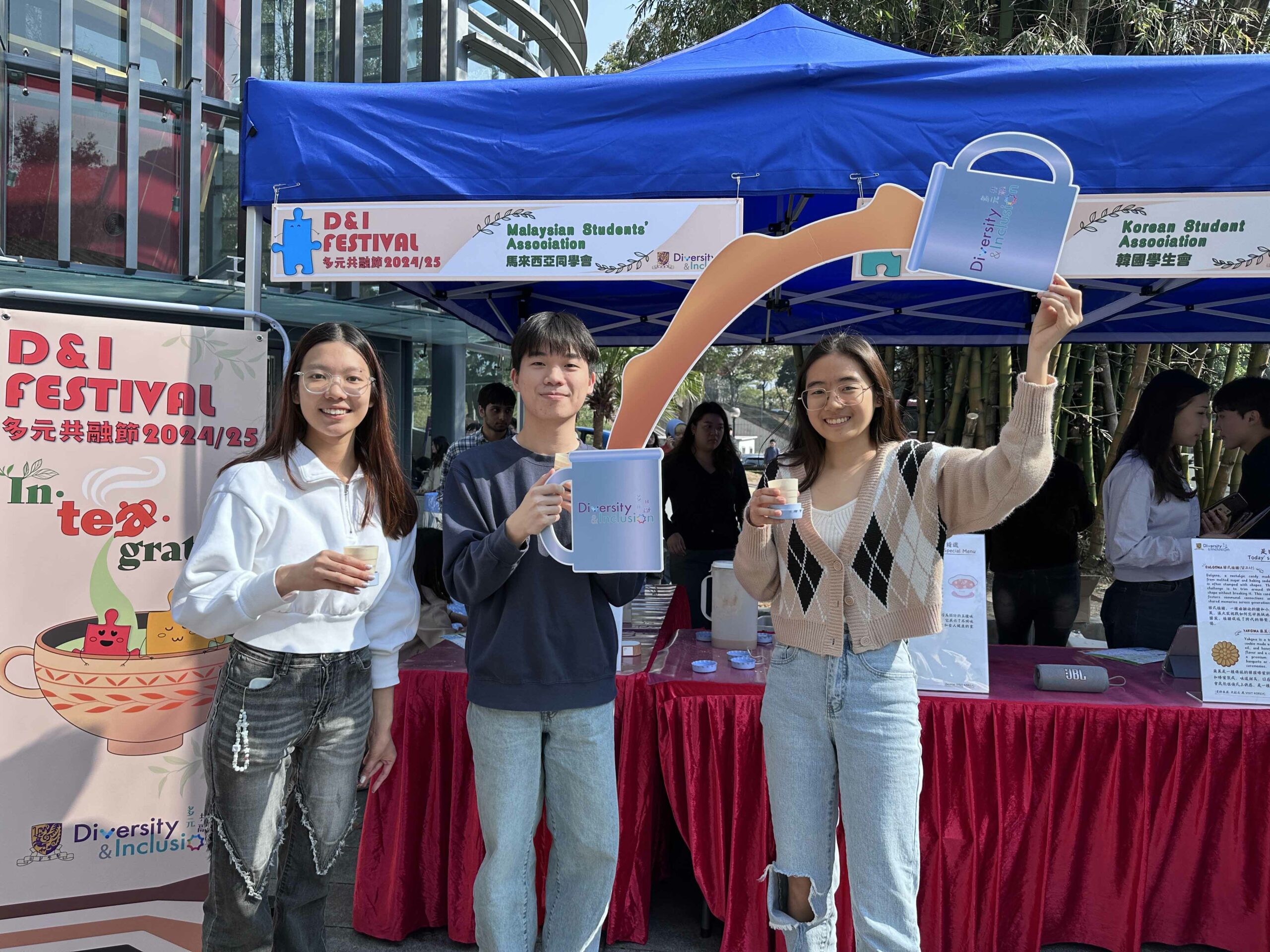 Three students from the Malaysia Students’ Association are smiling and taking photos with “Pull-tea” props under a blue tent with a "D&I Festival" banner.