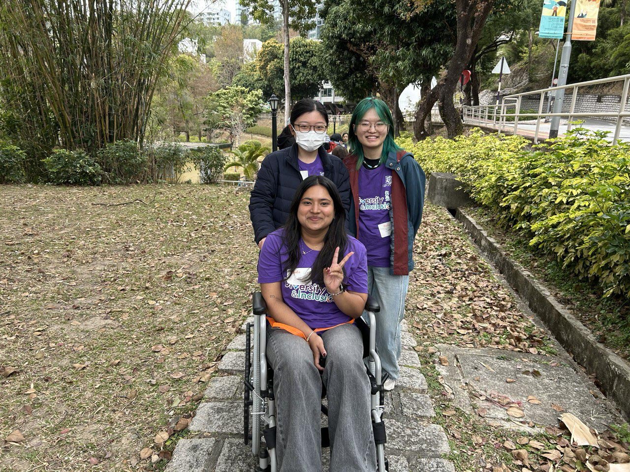 A D&I ambassador is seated in a wheelchair while two other ambassadors stand beside them, acting as caregivers and assisting with mobility.