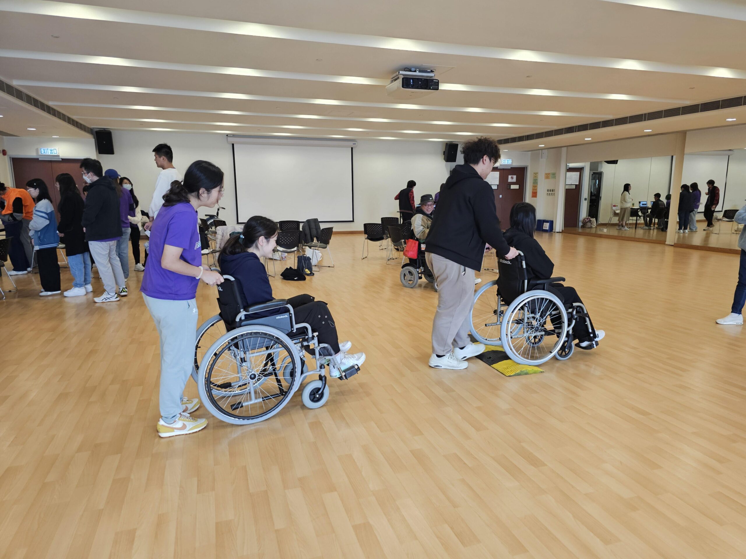 Two D&I ambassadors are seated in wheelchairs while two other ambassadors stand behind them, acting as caregivers and providing support.